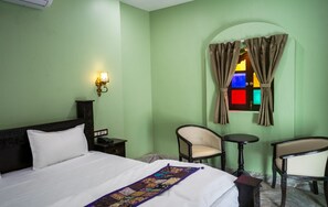 A hotel room with a bed, two chairs, a small table, a nightstand, a wall-mounted lamp, and a window with curtains.