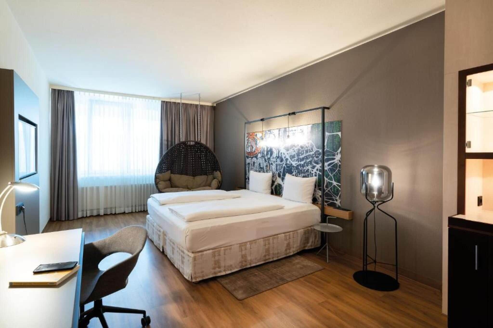 Une chambre d’hôtel moderne avec un grand lit, un bureau avec une chaise, un fauteuil suspendu et une lampe sur pied.