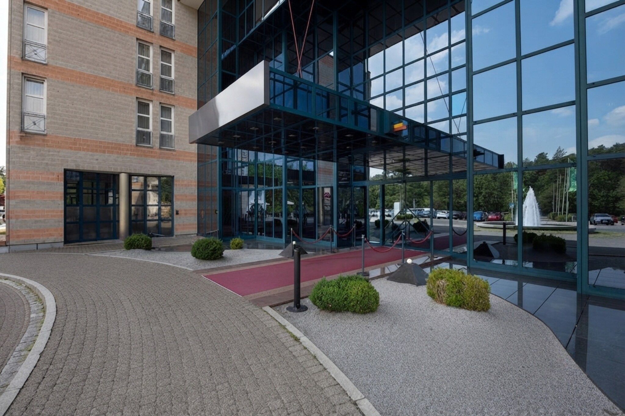 Ein modernes Gebäude mit Glasfassade, einem Ziegelunterbau und einem gepflasterten Eingangsbereich mit rotem Teppich und kleinen Büschen.