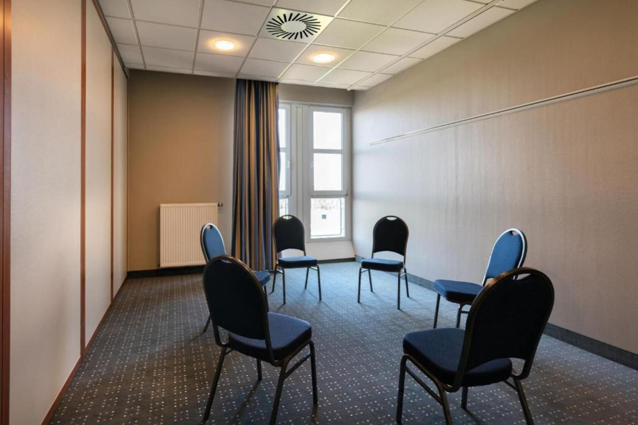 A conference room with a white ceiling, a radiator, and a window with curtains. There are four chairs arranged around a table.