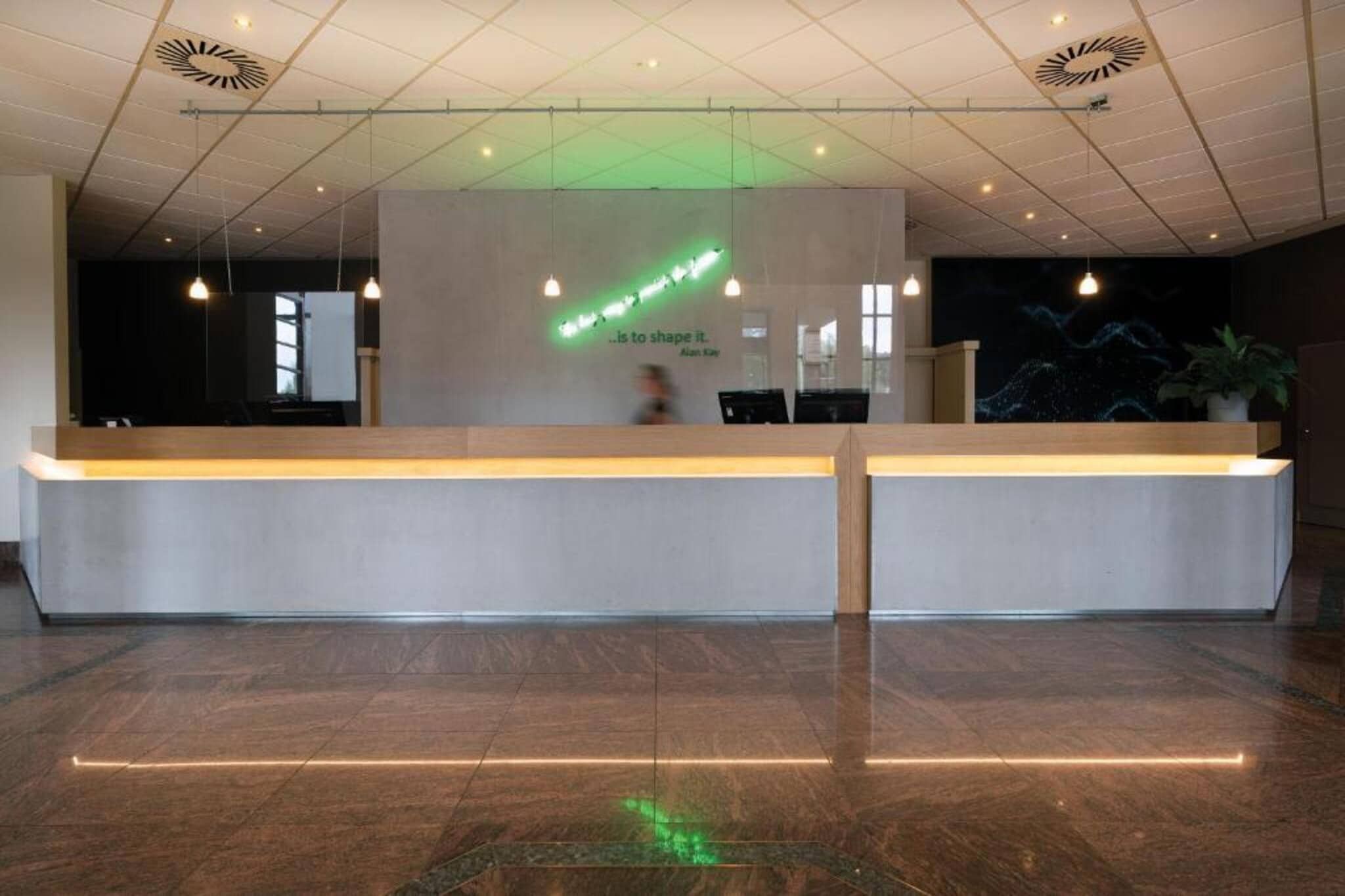 Un comptoir d’accueil au design moderne, une installation lumineuse verte sur le mur et une citation d’Alan Kay.