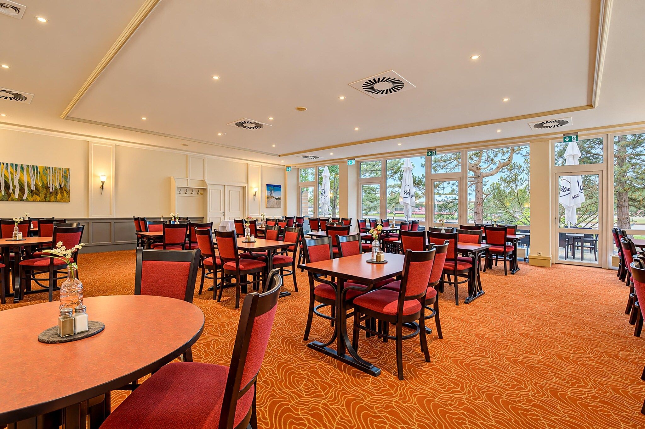 A hotel dining area with multiple tables and chairs, large windows, and a carpeted floor.