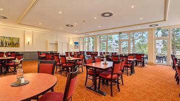 A hotel dining area with multiple tables and chairs, large windows, and a carpeted floor.