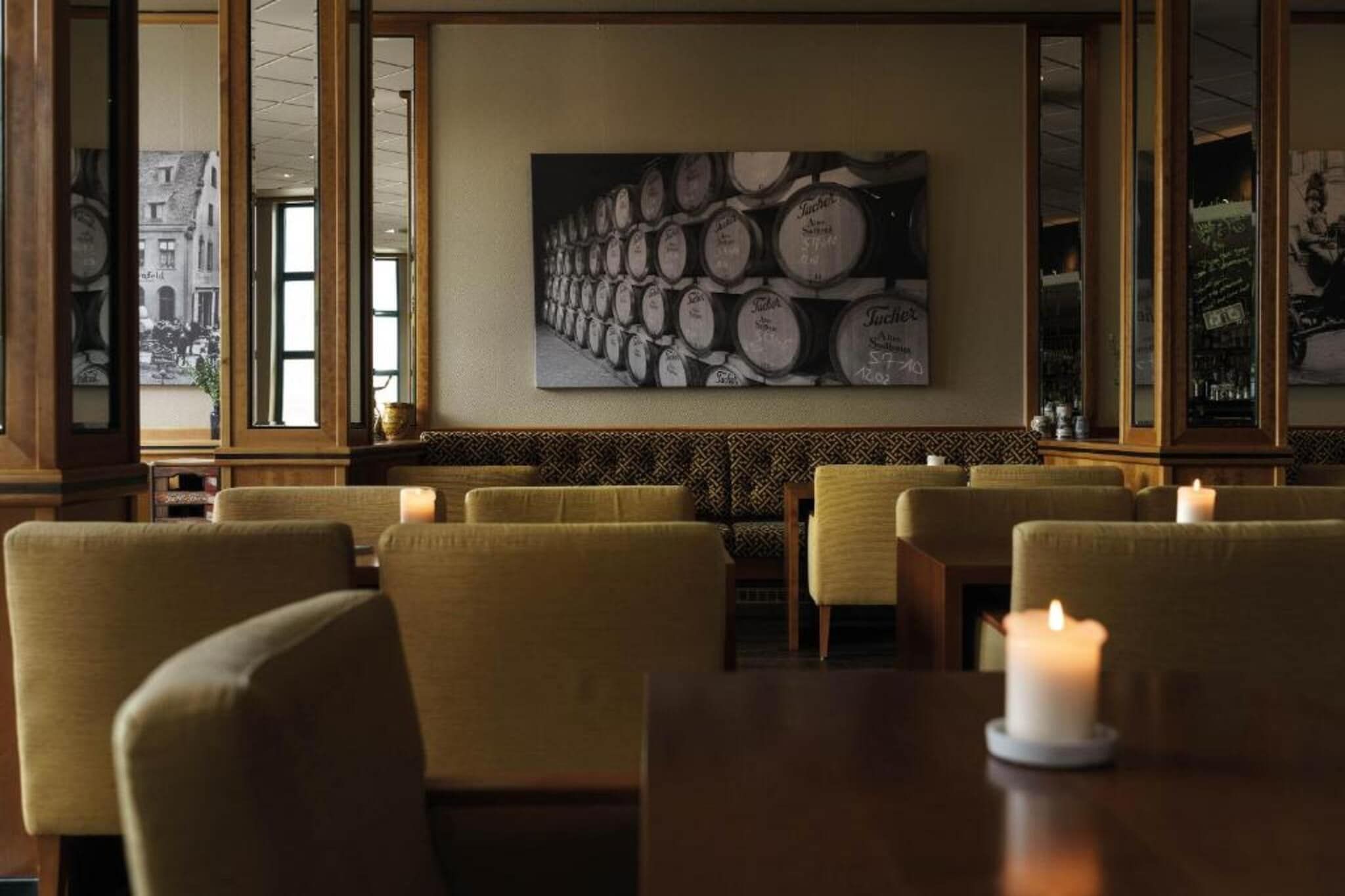 Een restaurantinterieur met een rij barkrukken, een wijnvattenwand en een kaars op de tafel.