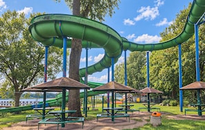 A park with a green water slide, picnic tables, and a large tree.