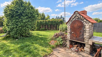 Une cheminée extérieure en pierre, dotée d’une porte en métal, entourée d’un jardin bien entretenu avec un grand arbre et un banc.
