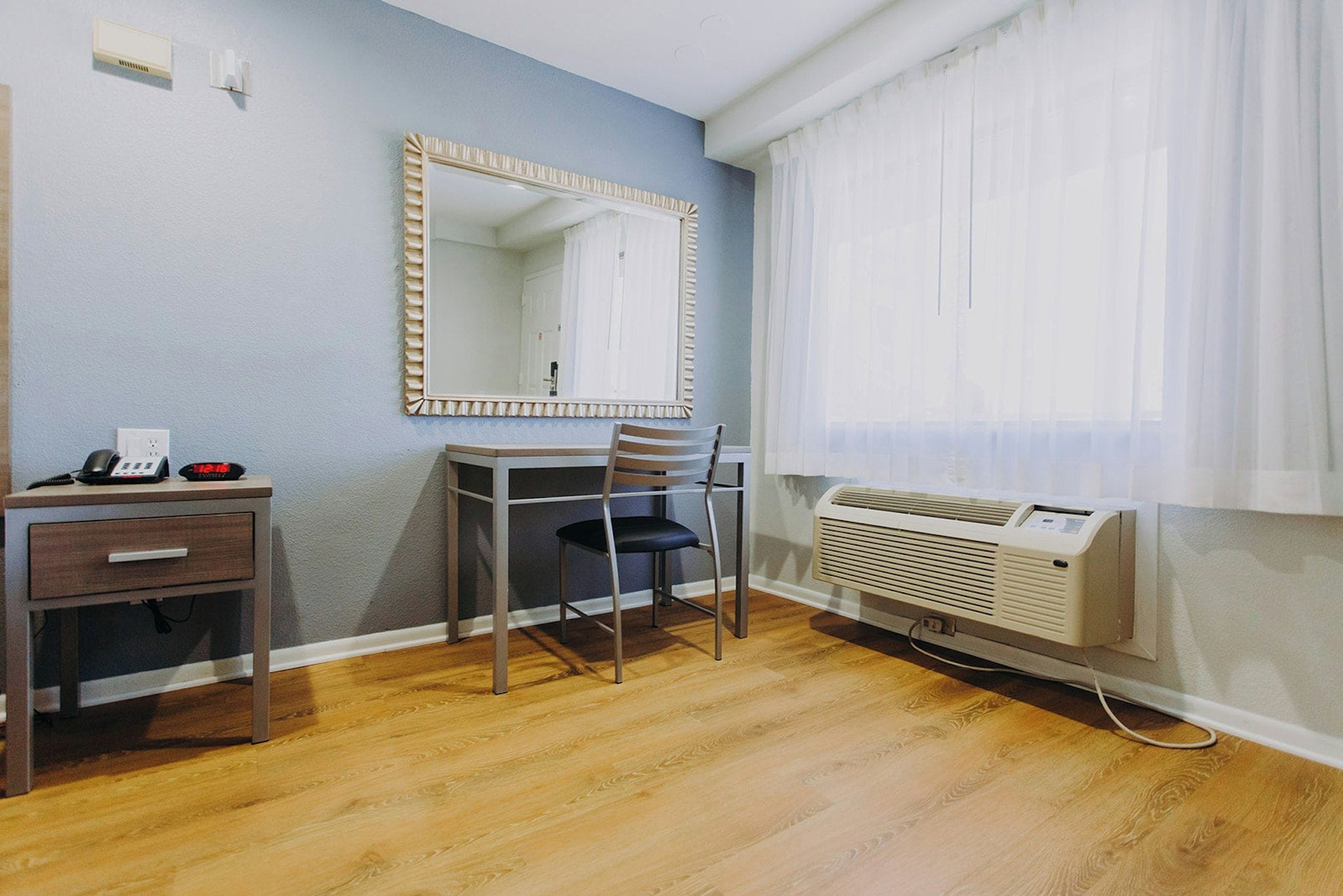 A room with a desk, chair, mirror, and a window with curtains.