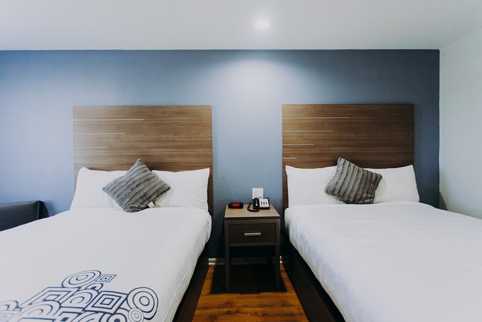 A hotel room with two beds, a nightstand with a phone and a clock, and a wooden headboard.