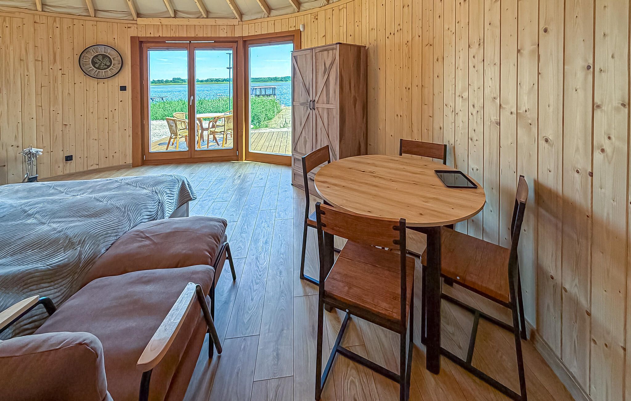Une pièce comprenant un lit, une table ronde en bois avec des chaises, une armoire et une vue sur une étendue d’eau, visible à travers des portes vitrées.