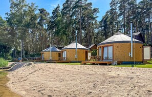 Alojamiento tipo yurta con exterior de madera y techo cónico, ubicado en una zona arenosa con un bosque de fondo.
