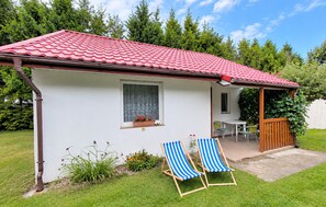 A small house with a red tiled roof, a window with a curtain, a patio with a table and chairs, and a garden with plants and two striped lounge chairs. - 2 bedroom beautiful home in Przewloka (Przewloka)