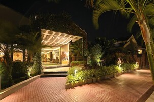 A well-lit building with a covered entrance, surrounded by lush greenery and a paved pathway.