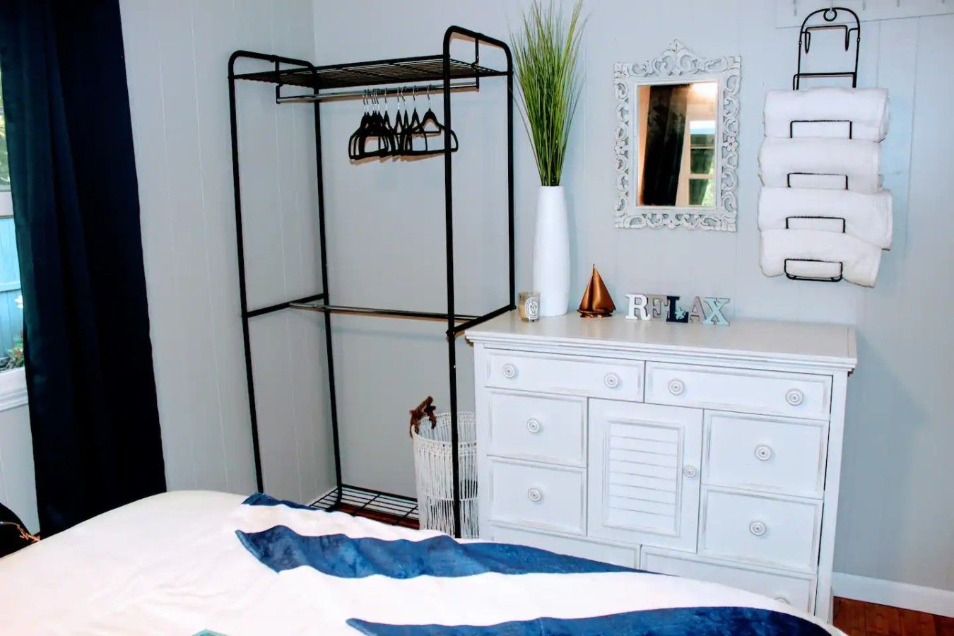 Une chambre à coucher comprenant un lit, une commode blanche, un étendoir métallique, un miroir et un porte-serviettes fixé au mur.