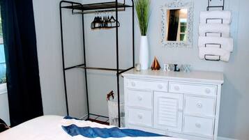 Une chambre à coucher comprenant un lit, une commode blanche, un étendoir métallique, un miroir et un porte-serviettes fixé au mur.