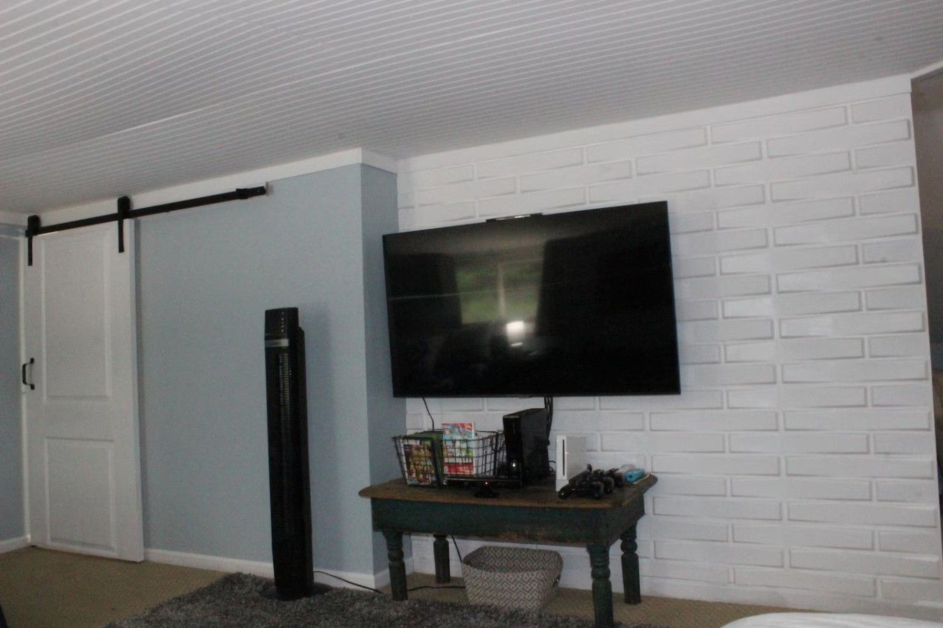 A room with a flat-screen TV mounted on a brick-patterned wall, a sliding door, a tall black speaker, and a small table with a basket.