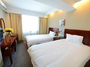 Ein Hotelzimmer mit zwei Betten, einem Schreibtisch mit Computer, einer Vase mit Blumen und einem Fenster mit Vorhängen.