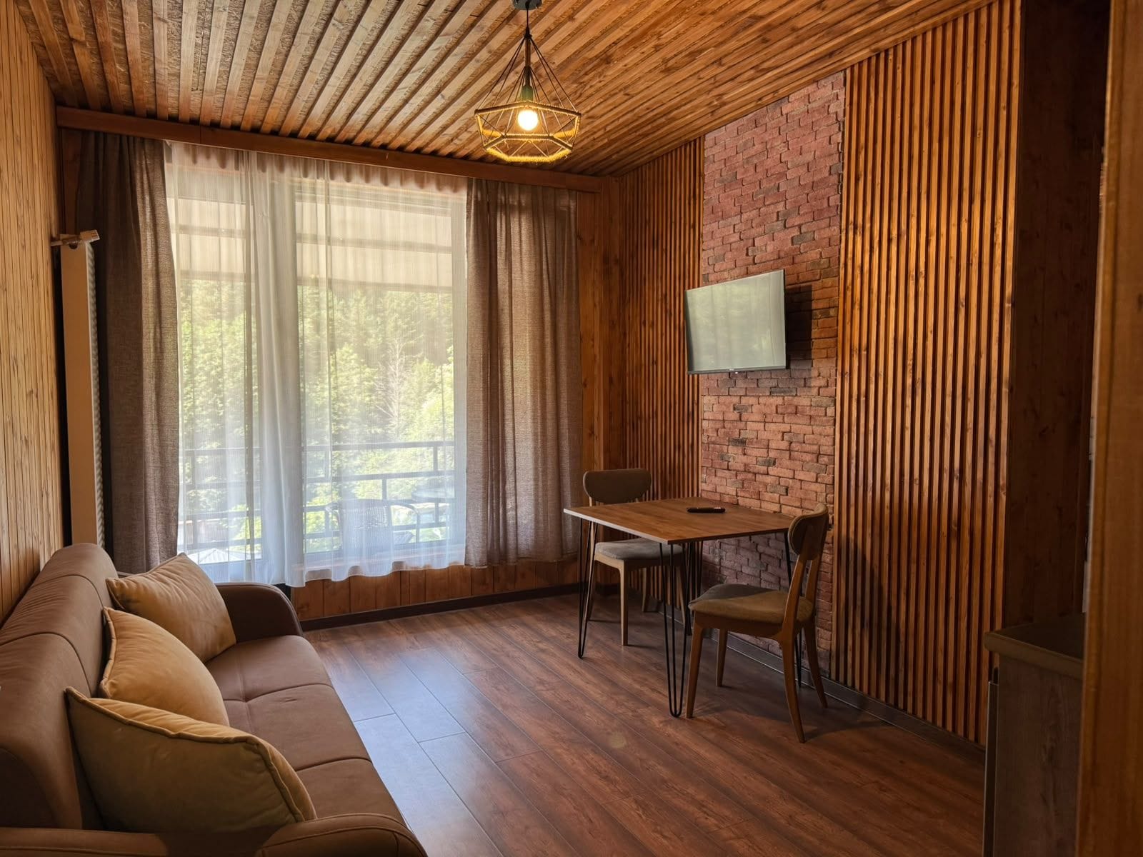 A cozy room with a wooden ceiling, a brick wall, a wooden floor, a sofa, a dining table with chairs, and a flat-screen TV mounted on the wall.