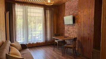 A cozy room with a wooden ceiling, a brick wall, a wooden floor, a sofa, a dining table with chairs, and a flat-screen TV mounted on the wall.