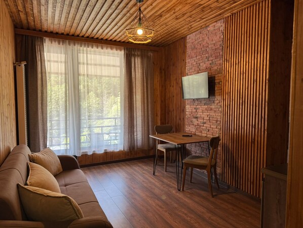 A cozy room with a wooden ceiling, a brick wall, a wooden floor, a sofa, a dining table with chairs, and a flat-screen TV mounted on the wall.