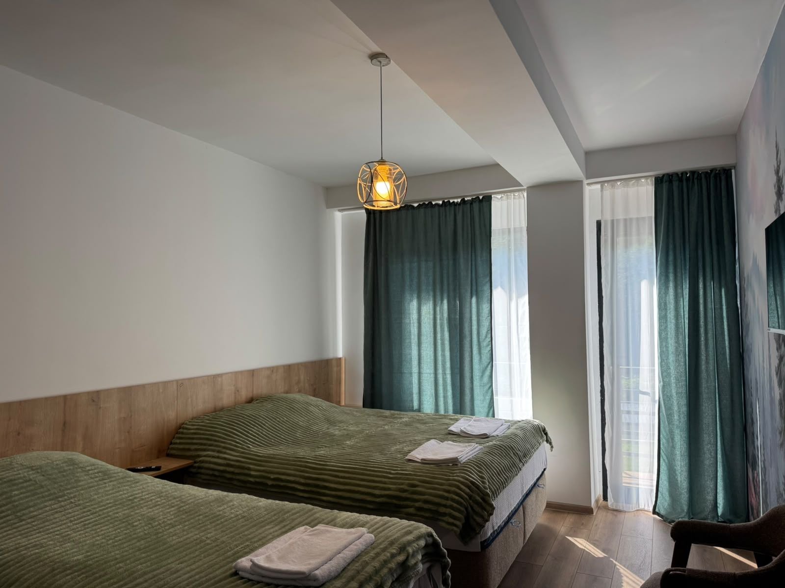 A hotel room with two beds, a wooden headboard, a hanging light fixture, and a view of the outdoors through sheer curtains.