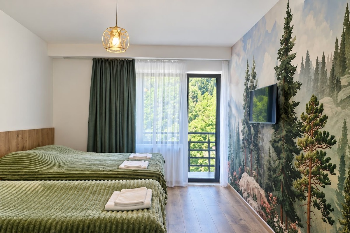 Un dormitorio con dos camas, un ventanal con cortinas, un cuadro de árboles y un televisor montado.