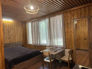 A room with a bed, a table with chairs, a hanging light fixture, and a window with curtains.