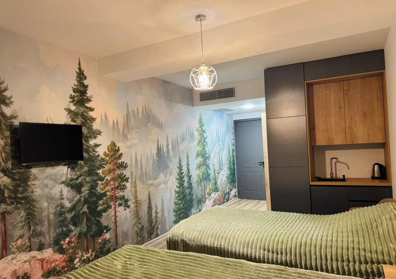A bedroom with a forest mural, a bed with a striped bedspread, a flat-screen TV, a wooden cabinet, and a pendant light.