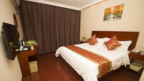 A hotel room with a large bed, a wooden headboard, a nightstand, a desk with a computer and plants, a large window with curtains, and a painting on the wall. - Greentree Inn Nantong Qidong Lvsi Harbour Express Hotel (Nantong)