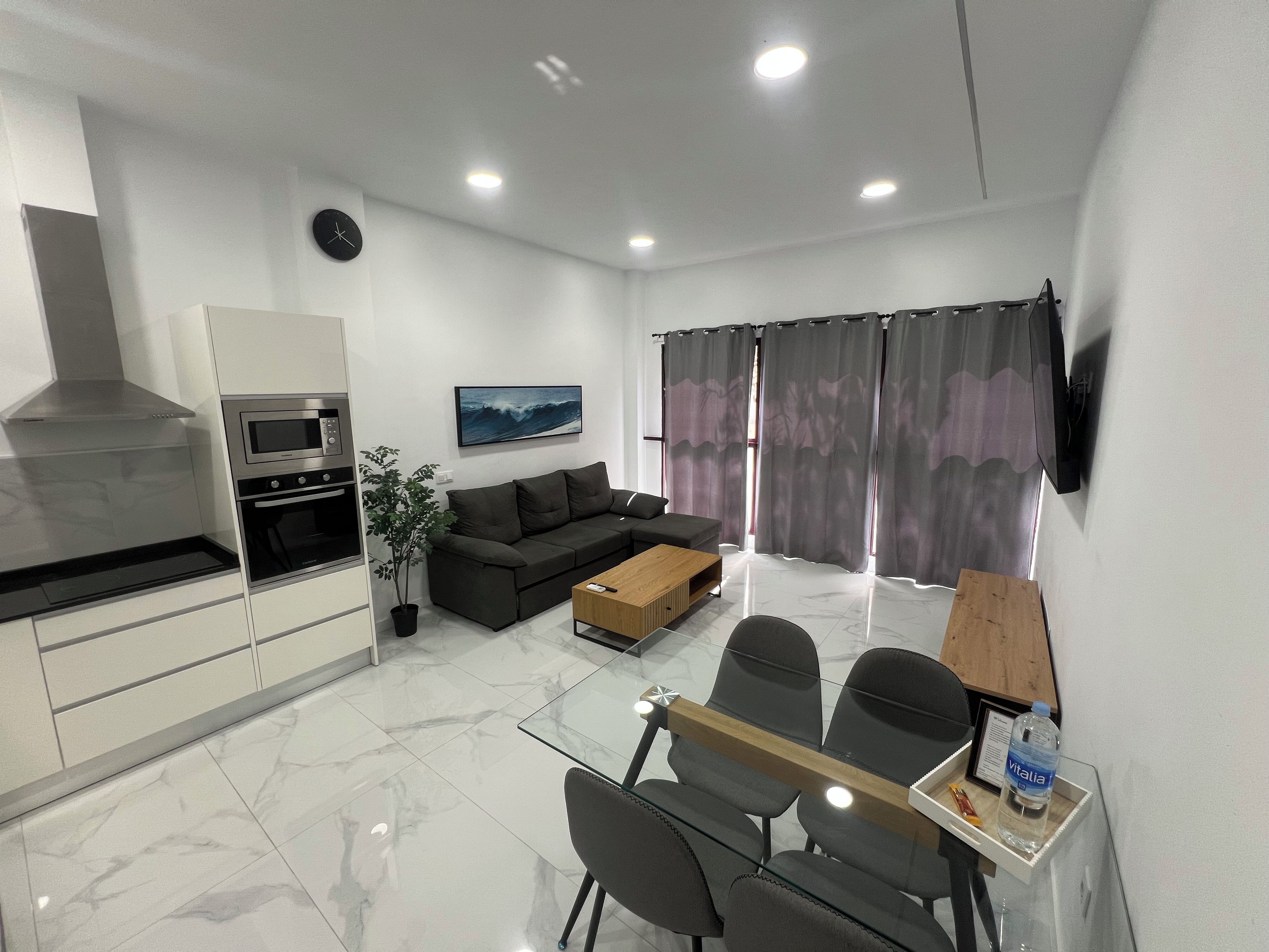 A modern kitchen with a built-in oven and microwave, a dining area with a glass table and chairs, a sofa, and a TV mounted on the wall.