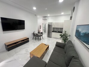 Ein modernes Wohnzimmer mit einem Flachbildfernseher, einem Holztisch, grauen Sofas und einem Essbereich mit einem Glastisch und Stühlen.
