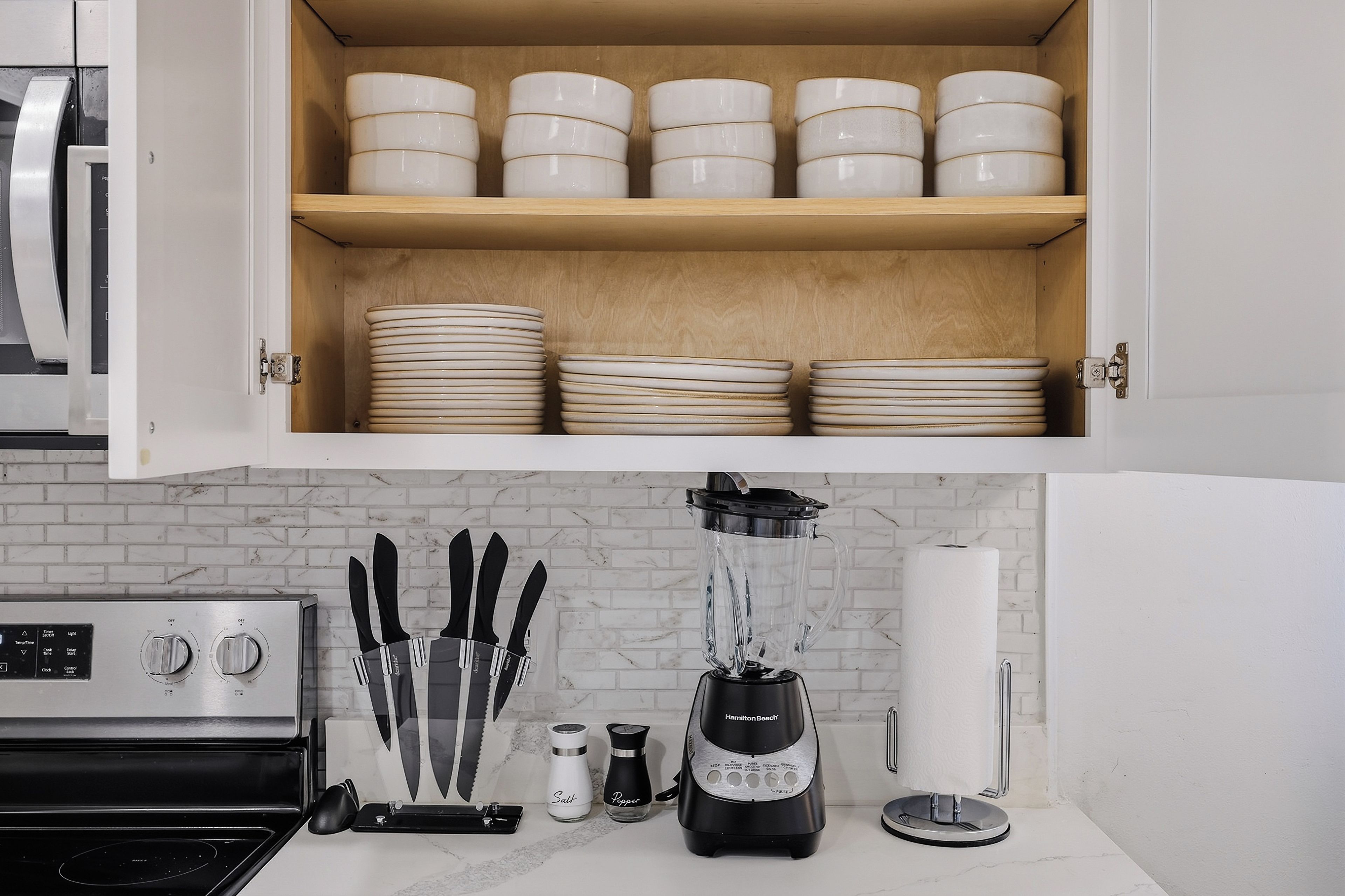 Una cocina con un mueble de madera que contiene platos y tazones apilados. Abajo, hay una encimera con una licuadora, un bloque de cuchillos, saleros y pimenteros, y un dispensador de toallas de papel.