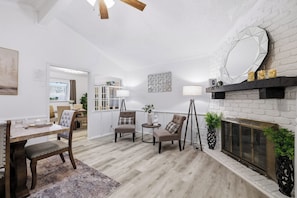 A modern living room with a fireplace, a dining area with a table and chairs, and a kitchen in the background.