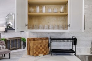 Una cocina con un armario de madera que contiene cristalería transparente, una tabla de cortar de madera y un estante de metal.