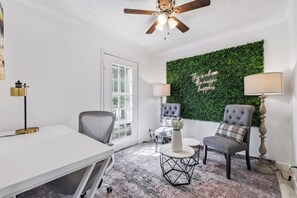 Una habitación con techo blanco, ventilador de techo, pared verde con una cita, dos sillas, una mesa y una lámpara de pie.