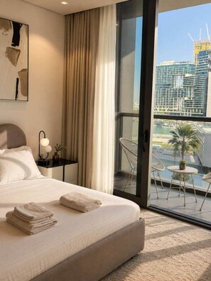 A hotel room with a bed, a nightstand with a lamp, a view of a cityscape through a large window, and a balcony with chairs and a small table. - New 1BR in Business Bay  Free Padel Court, Pool, Sauna (Dubai)