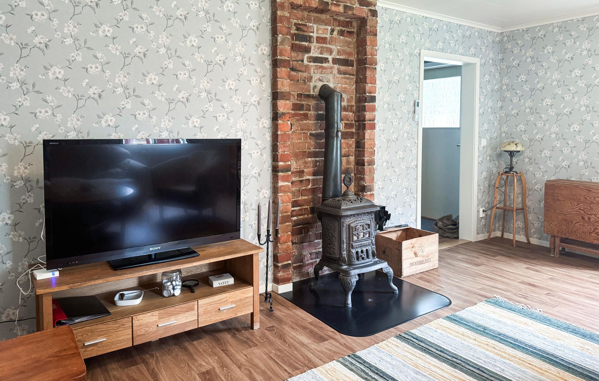 A living room with a brick fireplace, a flat-screen TV on a wooden stand, and a patterned wallpaper.