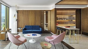 Una sala de estar moderna con un sofá azul, dos sillones rosa, una mesa de centro redonda y una zona de comedor con barra.