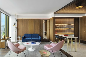 A modern living room with a blue sofa, two pink chairs, a round coffee table, and a dining area with a bar. - TWA Hotel (Jamaica)