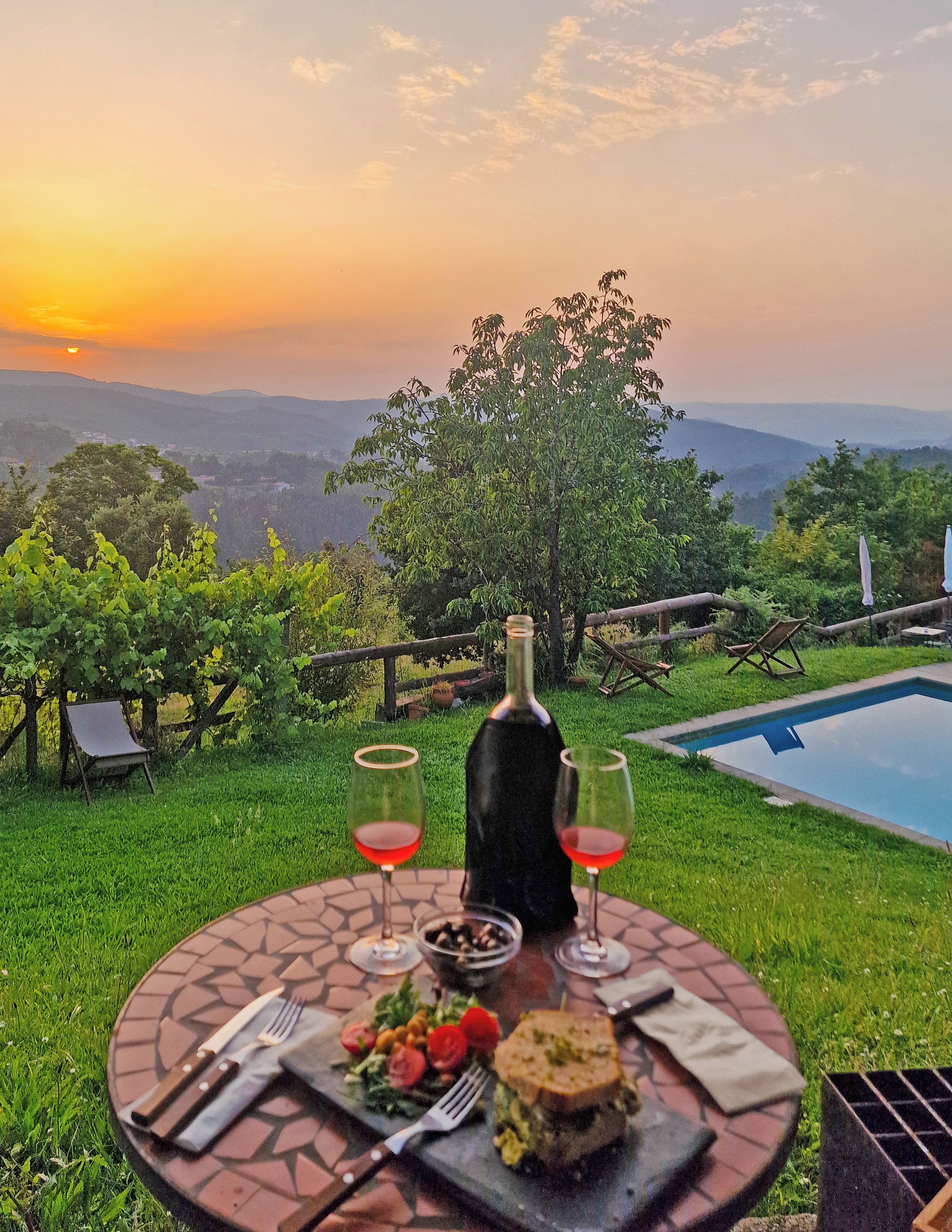 A table set outdoors with a bottle, glasses, and food, overlooking a pool and a scenic view.