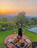 Mesa al aire libre con una botella, vasos y comida, con vistas a una piscina y un paisaje.
