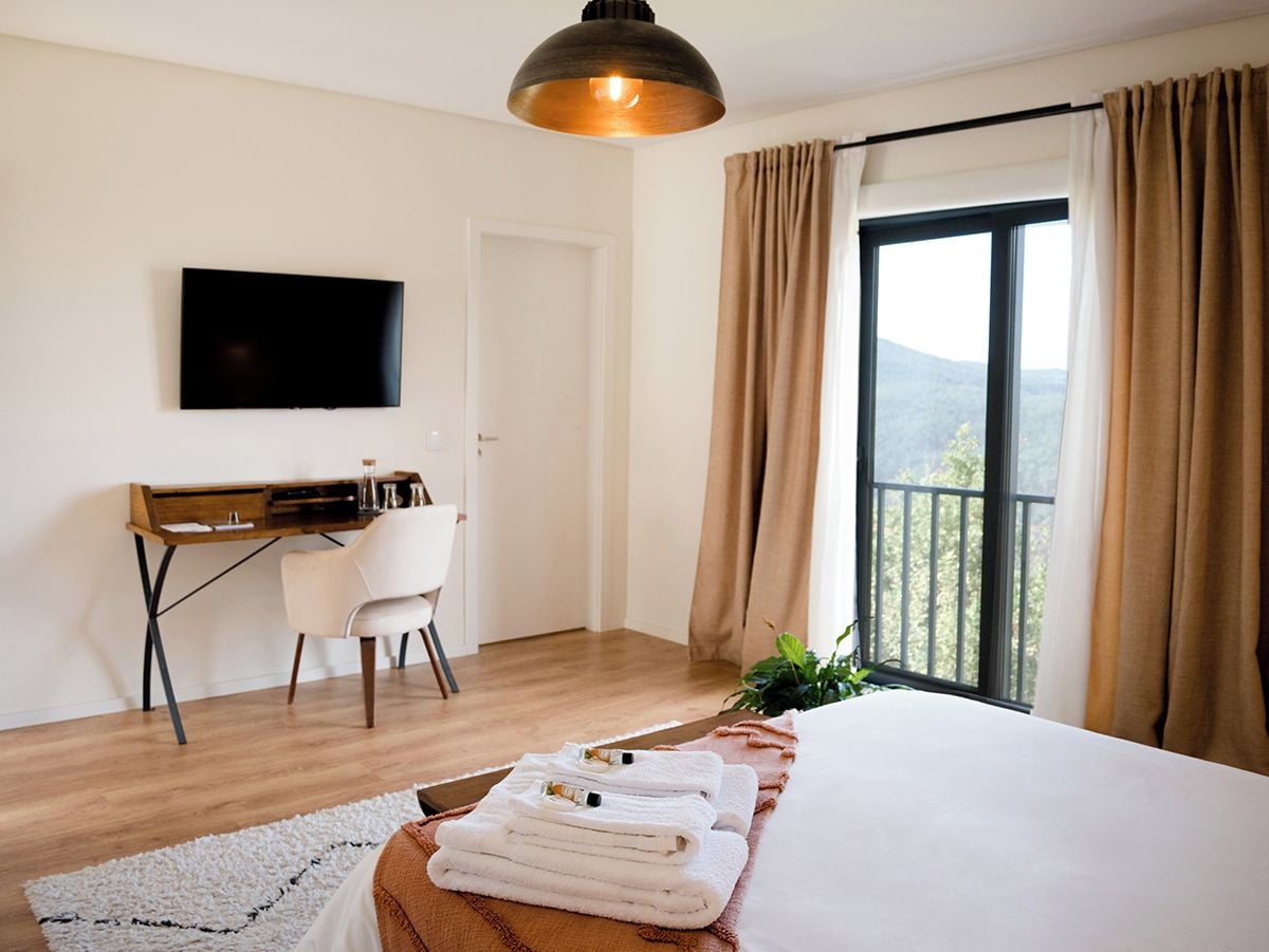 Una habitación de hotel con cama, escritorio, silla, televisor y balcón con cortinas.