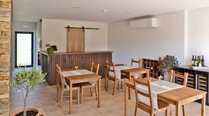 A restaurant with wooden tables and chairs, a bar area with wine bottles, and a small potted plant.