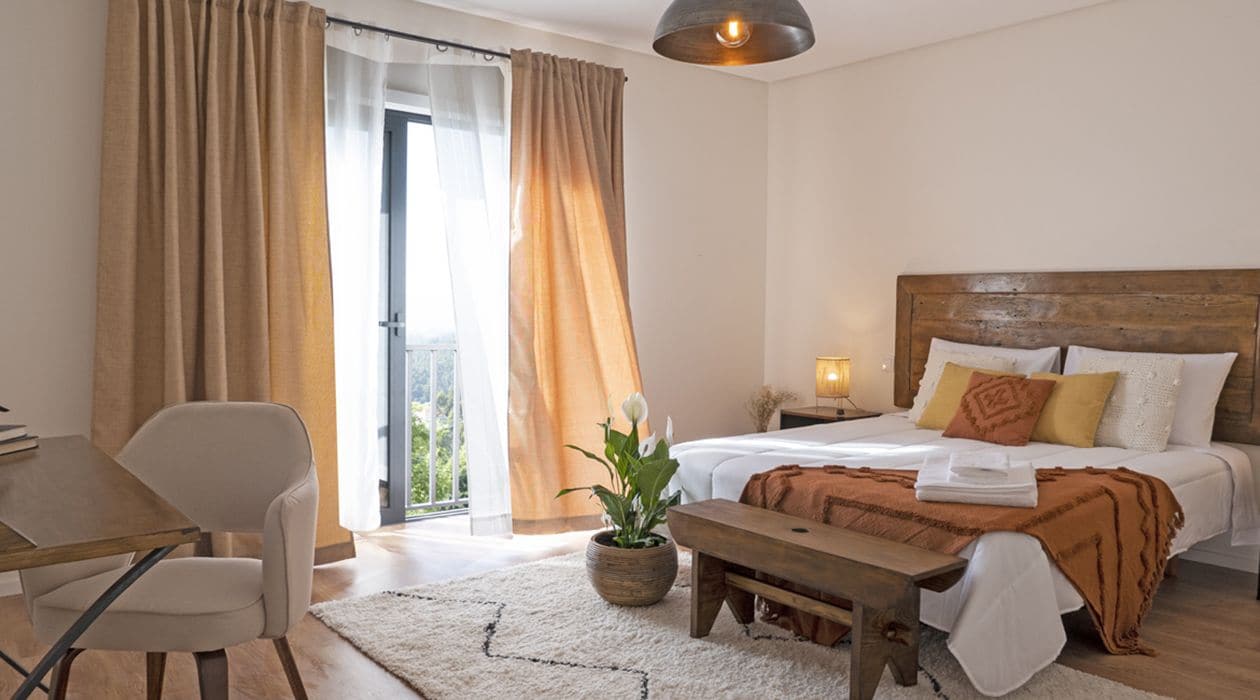 A hotel room with a bed, a desk, a chair, a potted plant, and a view of the outdoors.