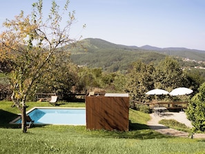 A swimming pool with a wooden fence, lounge chairs, and umbrellas in a backyard with a view of hills.