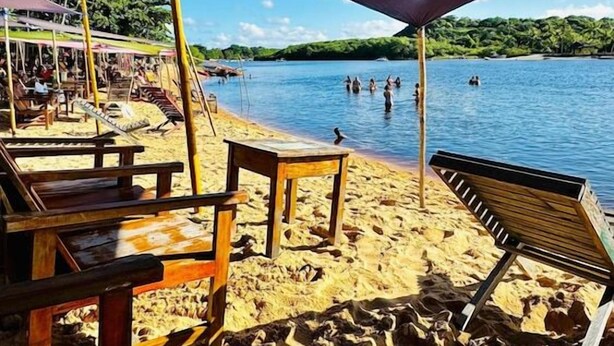 A beach with wooden lounge chairs and a shaded area, overlooking a calm body of water with people swimming. - Pousada Aquarela Caraiva (Porto Seguro)