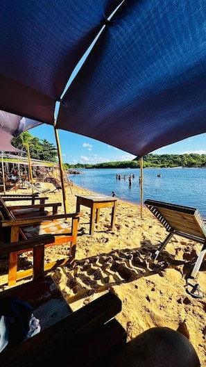 A beach with wooden lounge chairs and a shaded area, overlooking a calm body of water with people swimming. - Pousada Aquarela Caraiva (Porto Seguro)