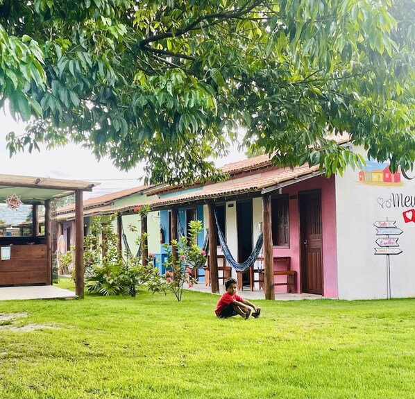 A building with a porch, a hammock, and a child sitting on the grass. - Pousada Aquarela Caraiva (Porto Seguro)