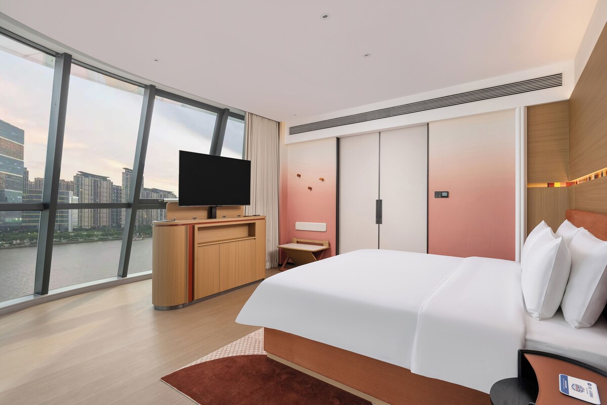 A modern hotel room with a large bed, a desk, a television, and a view of the cityscape.