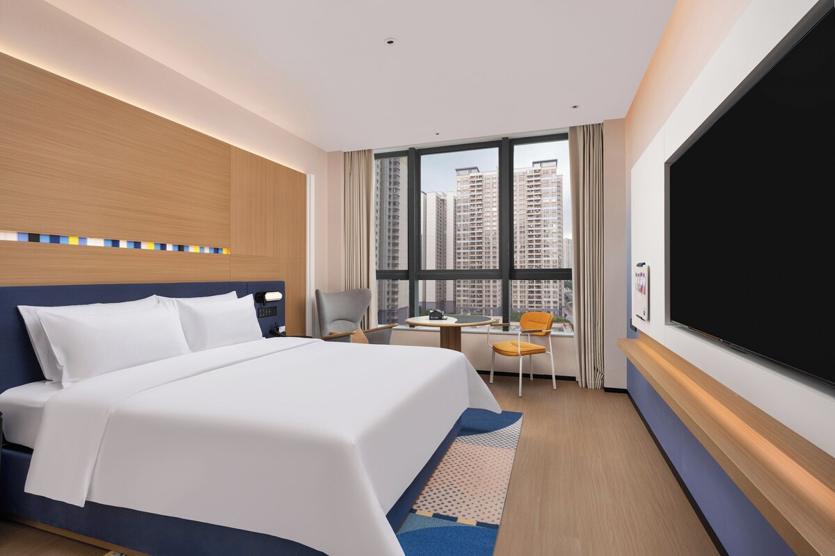 A hotel room with a large bed, a desk, a chair, a TV, and a view of tall buildings.