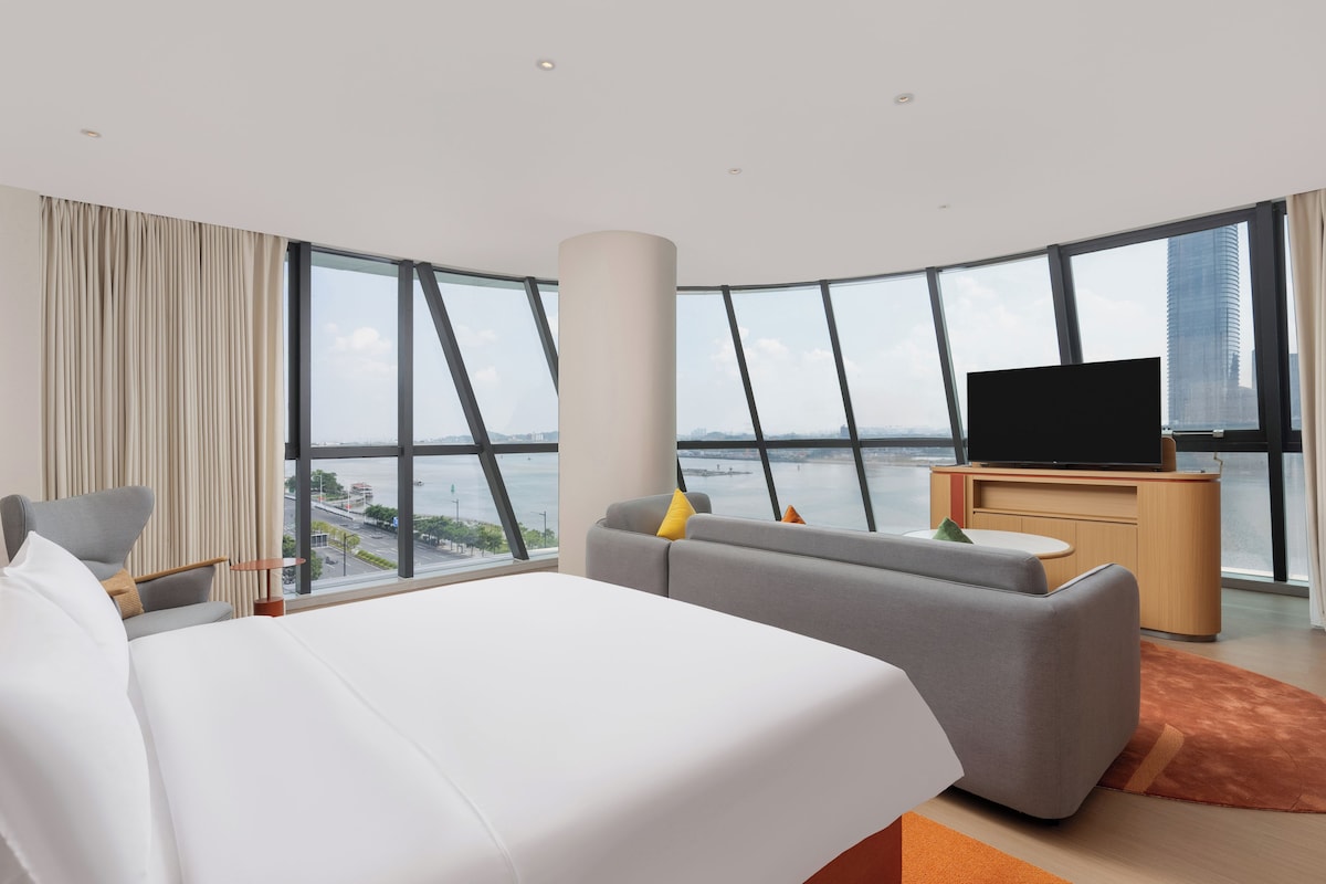 A modern hotel room with a large bed, a sofa, a TV on a wooden cabinet, and a view of a body of water through large windows.